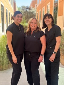 Three women dressed in black scrubs are standing together outdoors, smiling and posing for a photo. The setting appears to be near a building with greenery in the background, suggesting a professional or healthcare environment.