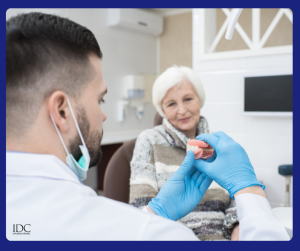 Dentist in mask and blue gloves shows a dental model or impression to an elderly female patient seated in chair; clinical setting with “IDC” watermark.