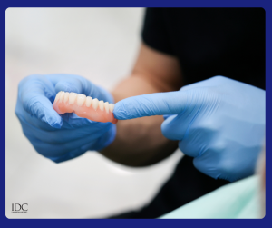 Gloved hands holding a pink-acrylic partial denture; index finger points to posterior teeth, highlighting fit or design
