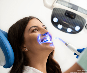 Patient in dental chair receiving in-office whitening; blue LED light activated, clinician’s gloved hand holding wand